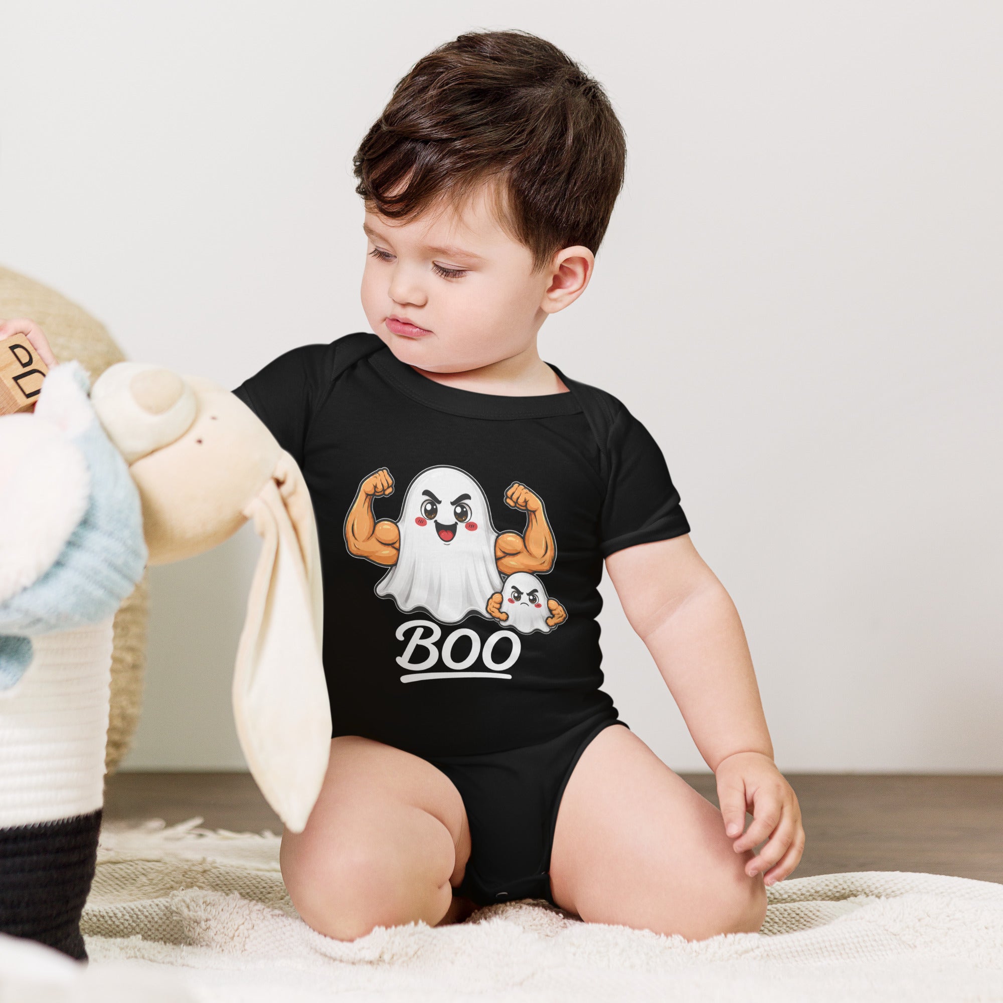 A toddler wearing the Boo Ghost Baby One Piece – Halloween Edition by Straight Face Originals, which features a strong, muscular cartoon ghost and “BOO” text, sits on the floor looking at a stuffed animal.