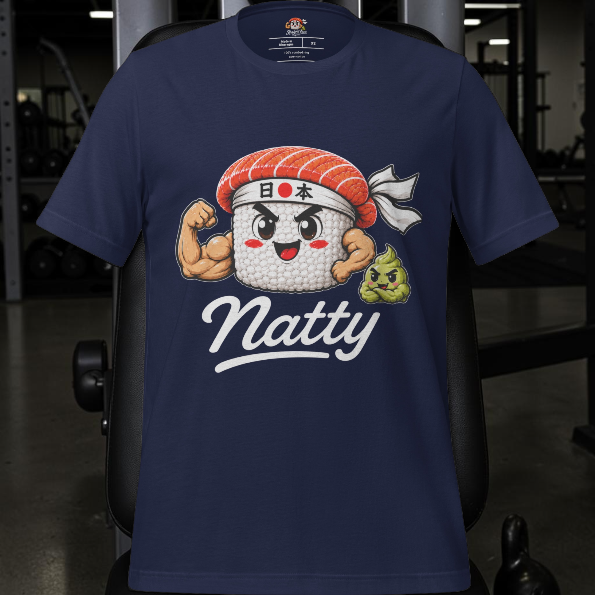 Introducing the Natty Tee - Japan Edition from Straight Face Originals: a navy blue t-shirt showcasing a cartoon sushi roll with muscular arms, a headband adorned with Japanese text, and a charming small green mascot. The word “Natty” is featured beneath the lively design. Perfect for gym enthusiasts seeking authentic clean carbs apparel with Japanese flair, this shirt is stylishly displayed here in a gym environment.